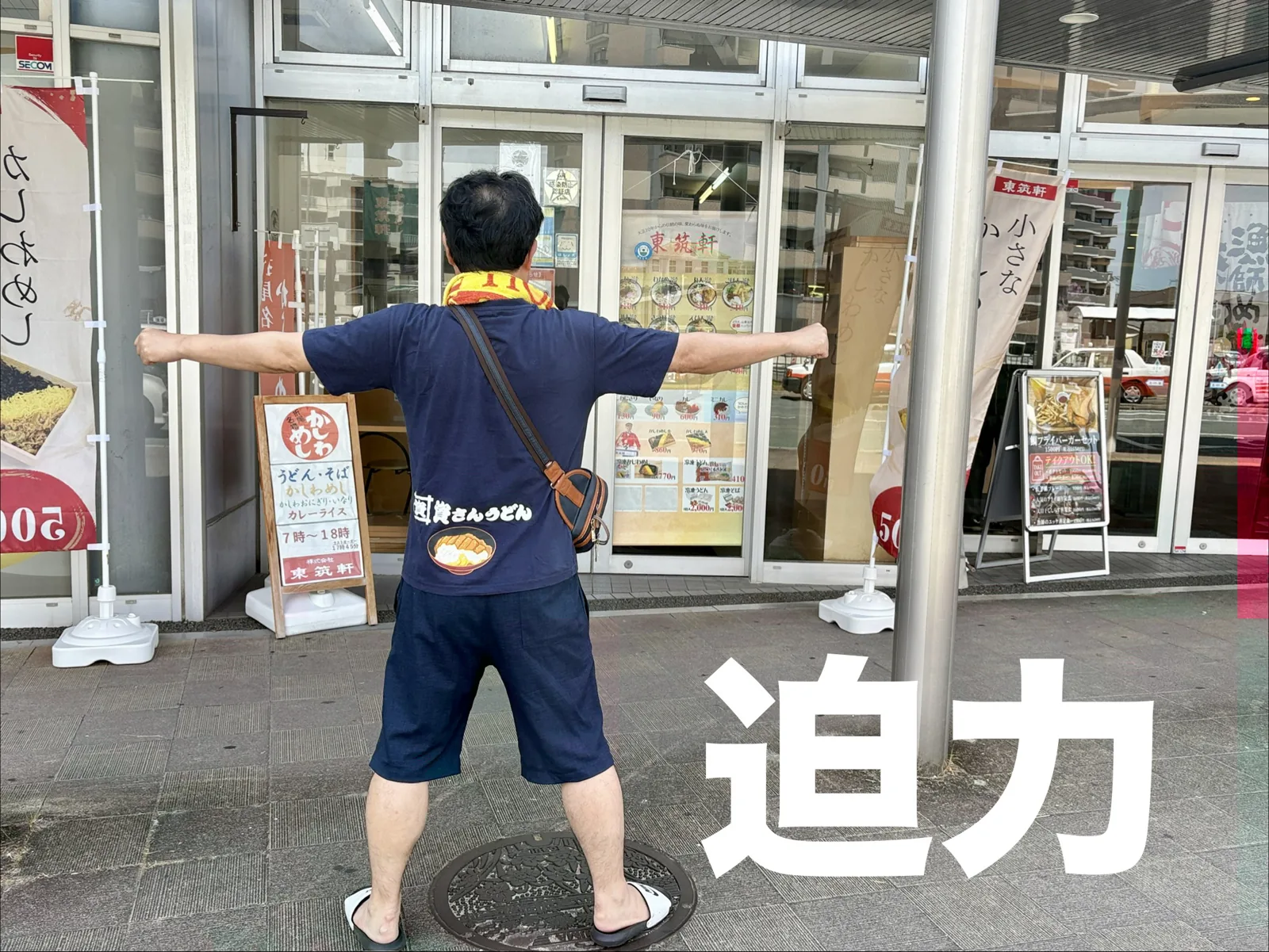 福間の店の前で迫力ポーズ。看板に「うどん・そば かしわめし カレーライス 7時〜18時」