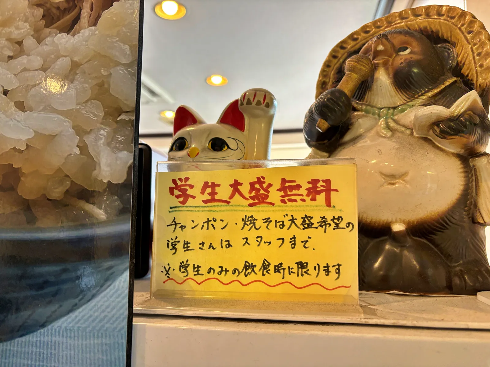 手書きの掲示「学生大盛無料」。招き猫とたぬきの置物の横に置かれている