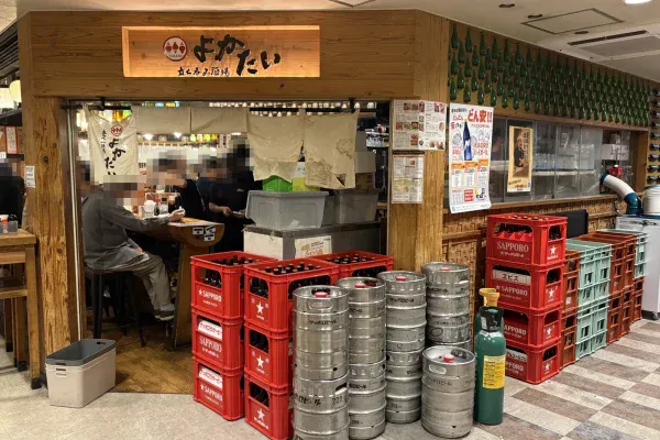 立ち呑み酒場よかたいの外観。木の看板とのれん、店頭に赤いサッポロのビールケースと銀色の樽が積み上がる