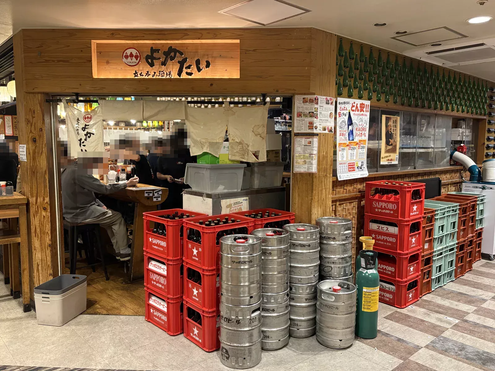 立ち呑み酒場よかたいの外観。木の看板とのれん、店頭に赤いサッポロのビールケースと銀色の樽が積み上がる