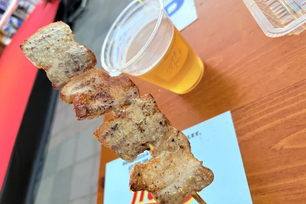炭火で焼いた豚バラ串を持ち上げ、奥にクロサキスイッチのロゴ入りカップのビール