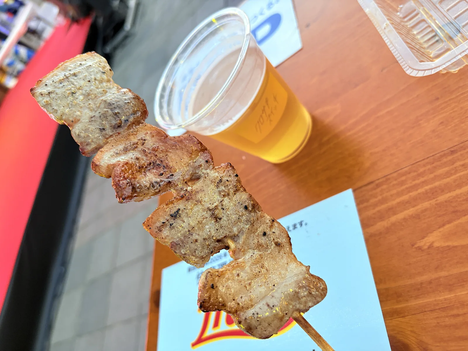 炭火で焼いた豚バラ串を持ち上げ、奥にクロサキスイッチのロゴ入りカップのビール