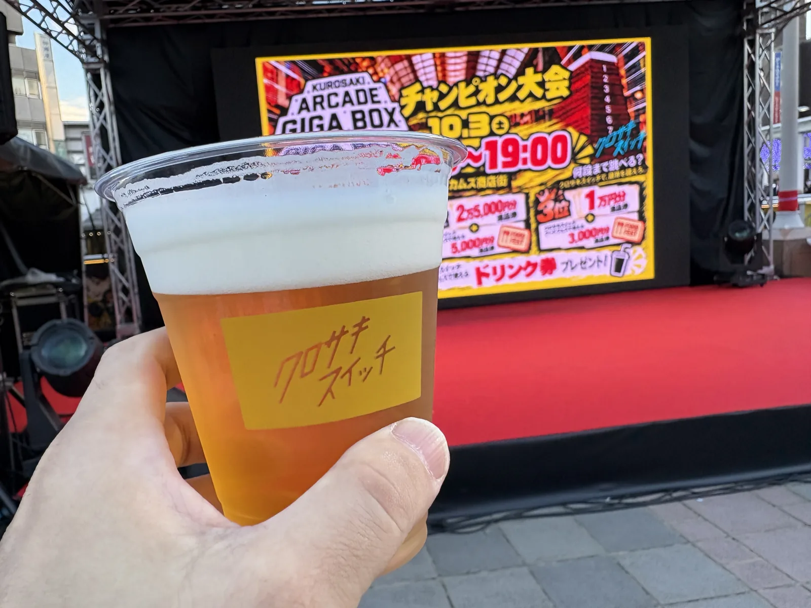 ステージ前でクロサキスイッチのロゴ入りカップの生ビールを掲げる。奥のモニターには跳び箱大会の告知