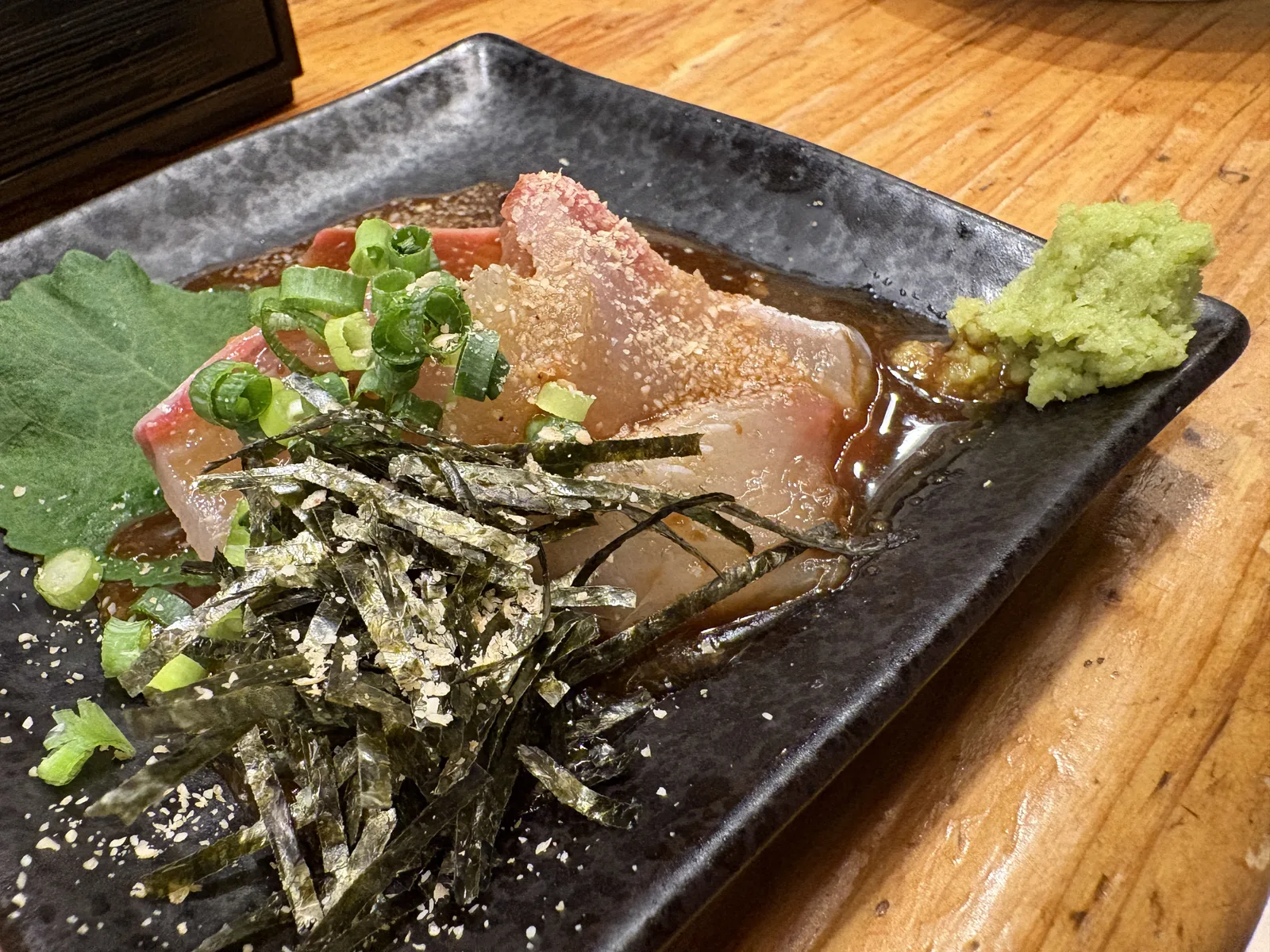ごまかんぱち。黒い角皿にカンパチの切り身、ごまだれ、刻みのり、ねぎ、大葉、わさび