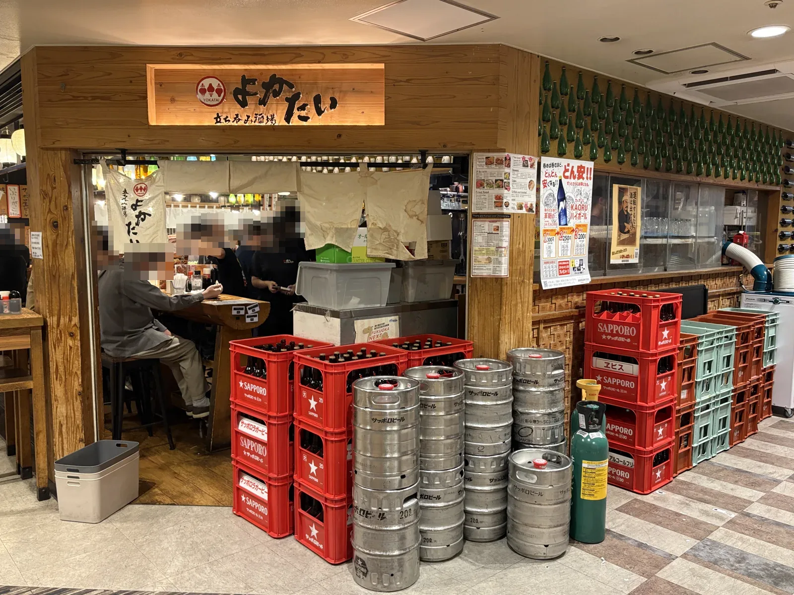立ち呑み酒場よかたいの外観。木の看板とのれん、店頭に赤いサッポロのビールケースと銀色の樽が積み上がる